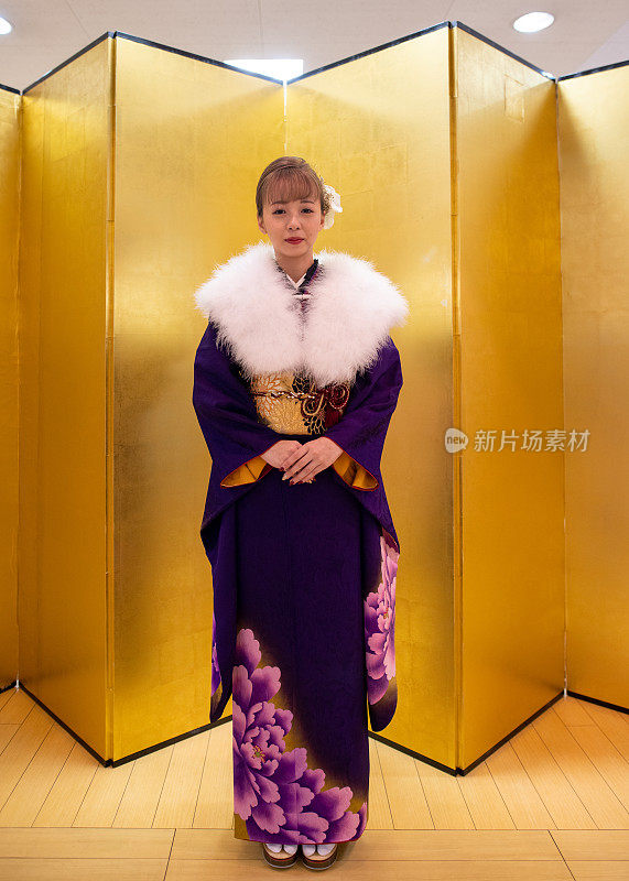 在“Seijin Shiki”成人礼上，年轻女子身穿Furisode和服，披着毛皮领披肩站在金色的Byobu折叠屏风前的全长肖像